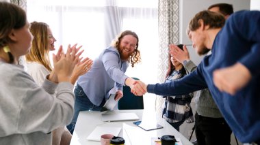 Şirket çalışanları başarılı bir işlemi kutluyorlar. Bir grup insan el sıkışıyor ve alkışlıyorlar. Sahne olumlu ve dostane. Takım çalışması.