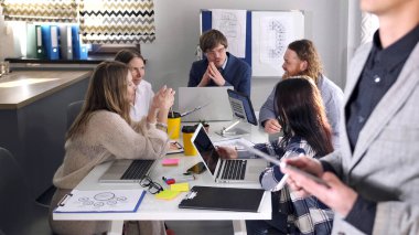 Bir grup insan bir masada oturuyor ve bir adam onların önünde duruyor. Bir adam elinde bir tablet tutuyor ve bir sunum yapıyor. Sahne profesyonel ve odaklı.