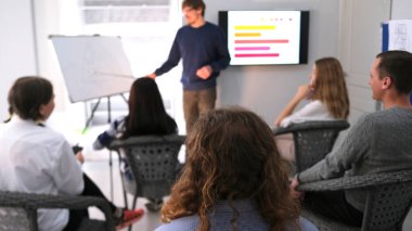 Genç bir stajyer meslektaşlarına sunum yapıyor. Birkaç kişi öğretim üyesinin önünde sandalyelerde oturuyor. Antrenörden iş eğitimi.