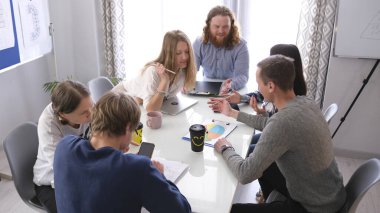 Bir grup insan bir masada oturuyor ve birlikte bir proje üzerinde çalışıyorlar. Herkesin kendi işiyle meşgul olduğu bir işbirliği ve konsantrasyon ortamı.