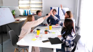 Yaratıcı iş ekibi ihaleyi kazanmayı kutluyor. Masadaki bir grup mutlu genç birbirine beşlik çakıyor. Resimde beş kişi var, ikisi kadın..