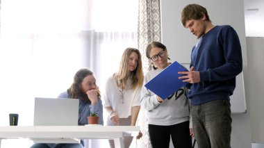 Meslektaşlar logo taslağını tartışıyorlar. Bir grup insan ellerinde mavi bir klasörle müdürün masasının etrafında duruyor. Sahne işbirlikçi ve odaklı.