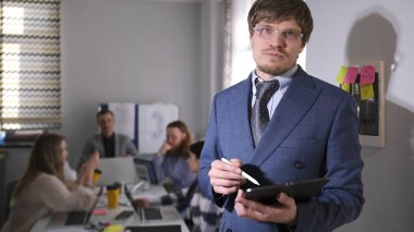 Başarılı, takım elbiseli ve gözlüklü bir adam bir grup insanın önünde duruyor. Elinde bir kalem ve tablet var. Sahne profesyonel ve ciddi.