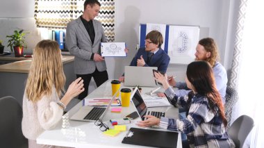 Kârlılık, yenilik ve şirketin yeniden yapılandırılmasının tartışılması. Bir grup insan bir masada oturuyor ve bir adam önlerinde duruyor ve bir sunum yapıyor.