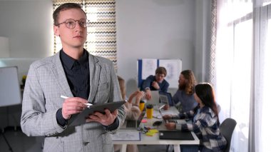 Takım elbiseli ve gözlüklü genç bir adam elinde tabletle bir grup insanın önünde duruyor. Bir çalışma toplantısı. Bir iş şirketinde sıradan bir gün.