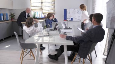 Bir grup iş adamı, ekonomi ve finans üzerine bir konferans salonunda bir masada oturuyor. Beyaz bir kurulun önünde elinde bir kalemle duran ve konferans veren bir kadın..