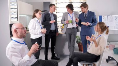 İşte öğle yemeği molası. Bir grup insan mutfakta toplandı, bazıları kahve içip salata yedi. Sohbet etmek için bir araya geldiklerinde ortam sakin ve dost canlısı görünüyor..