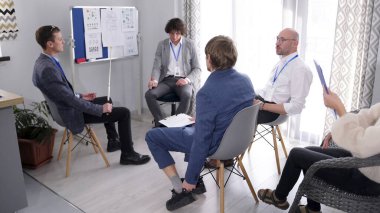 Danışmanlık şirketinin personeli gelecek sunum için hazırlanıyor. Bir grup insan ellerinde grafik ve diyagramlarla bir tahtanın etrafında oturuyorlar..