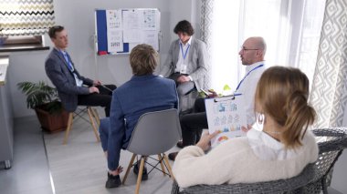 Bir iş şirketinde sabah brifingi. Bir grup insan bir konferans odasında sandalyelerde oturmuş sunum bekliyorlar. Sahne profesyonel ve odaklı.