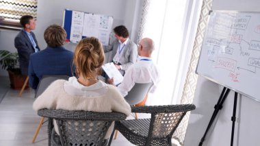 Şirketin finansörü bir sunum yapıyor. Bir grup iş adamı sandalyede oturup grafikler ve diyagramlarla dolu bir tahtaya bakıyor. Sahne odaklı ve ciddi.