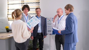 Şirket çalışanları grafikler ve diyagramlarla karatahtanın önünde neşeli bir şekilde sohbet ediyorlar. Bir kadın iş arkadaşlarına komik bir hikaye anlatır. Sunumu tartışıyorlar.