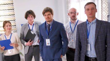 Takım elbiseli bir grup kadın ve erkek konferans odasında dikiliyorlar. Herkesin göğsünde isim kartı var ve bazılarının elinde klasörler var. Ortam profesyonel ve resmi.