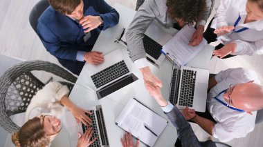 Önemli bir işlemi sonuçlandırma süreci, üst görüş. Bir grup işadamı dizüstü bilgisayar ve kağıtlarla aynı masada oturarak el sıkışıyor. Atmosfer profesyonel ve dost canlısı.