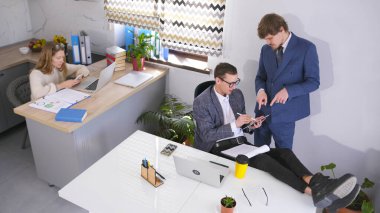 Şirket başkanı tablet için anlaşmayı imzalar. Firmanın avukatı patrona yeni bir müvekkil hakkında tavsiyede bulunuyor. Bir kadın sekreter iş adamlarının arkasında oturuyor..