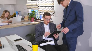 Şirketin müdürü kontratı imzalaması için patrona getirdi. Takım elbiseli ve gözlüklü bir girişimci tablet üzerinde elektronik bir anlaşma imzalıyor. Atmosfer profesyonel görünüyor.