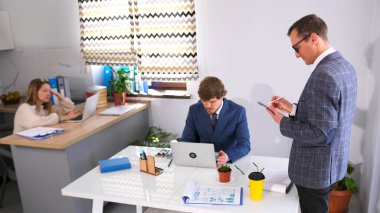 Genç, yakışıklı bir stajyer elinde bir tablet ve kalemle baş masasında duruyor ve talimatlarını yazıyor. Arkalarında bir kadın sekreter oturuyor. Atmosfer profesyonel ve odaklı.