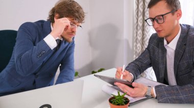 Gözlüklü iki genç girişimci bir masada oturuyorlar, biri tablete bir şey yazıyor. Başka bir işadamı tablete bakar ve içindekileri tartışır.