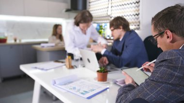 Bir grup ekonomist bir aile ofisinin mutfağında bir masada oturuyor ve bir proje üzerinde çalışıyor. Masanın üzerinde dizüstü bilgisayar ve tablet var, yanında da saksı bitkisi..