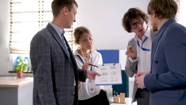 Bir adam elinde grafiklerle bir tablet tutuyor. Bir grup danışman onun yanında duruyor ve sunumun detaylarını tartışıyor. Sahne profesyonel ve odaklı.