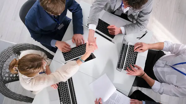 İş adamları dizüstü bilgisayarlarla dolu bir masada oturuyorlar. El sıkışıyorlar ve birlikte çalışıyorlar. İyi bir anlaşma. Selamlar. Toplantıda çalışma saatleri