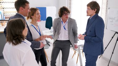 Şirketin genç kreatif direktörü astlarıyla iletişim kuruyor. Bir grup ofis çalışanı yarım daire şeklinde duruyor ve önemli bir projeyi tartışıyorlar..