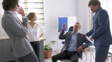 Danışmanlık şirketinin çalışanları toplantılar arasında birbirleriyle iletişim kurarlar. Bir grup erkek ve kadın bir konferans odasında duruyorlar. Sahne tasasız ve rahat.