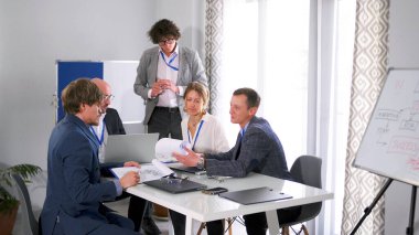 Bir grup insan konferans odasında bir masada oturuyor. Bir projeyi tartışıyorlar ve önlerinde diyagramlar olan bir yazı tahtası var. Sahne profesyonel ve odaklı.