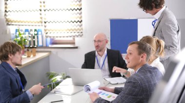 Bir grup adam konferans odasında bir masada oturmuş iş meselelerini tartışıyorlar. Atmosfer profesyonel ve odaklı, herkes sohbete dahil..