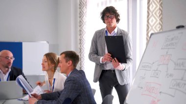 Yönetici raporunu toplantıda bitirdi ve masadaki yerine gitti. Gözlüklü, elinde klasör olan pozitif bir adam beyaz bir panonun önünde duruyor. Elinde bir iş projesi çizelgesi var..