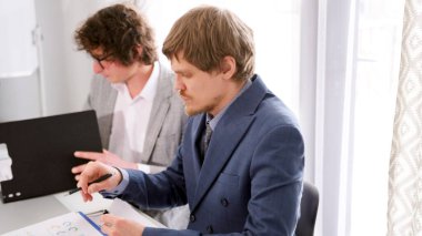 İki genç profesyonel avukat ortak bir dava üzerinde çalışıyor. İki adam bir masada oturuyor, biri kalemle bir şey yazıyor. Soldaki adam gözlük takıyor ve sağdaki adam da takım elbise giyiyor..