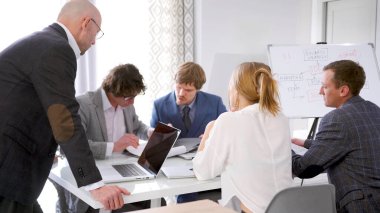 Şirketin müdürü ayağa kalktı ve masaya yaslandı. Bir grup ofis çalışanı toplantı odasında beyaz bir masada, arkasında bir iş fikrinin diyagramı olan bir tahtayla çalışıyor. Sabah toplantısı. Brifing..