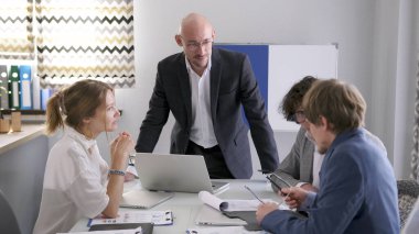 Yöneticiler Kurulu. Birkaç kişi bir dizüstü bilgisayar ve kağıtlarla bir masada oturuyor ve önemli bir projeyi tartışıyorlar. Takım elbiseli cana yakın, kendinden emin bir adam önlerinde duruyor ve talimatlar veriyor..