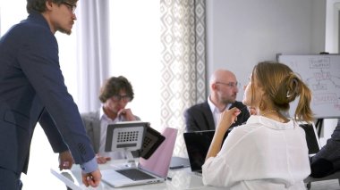 Bir iş şirketinde ciddi bir brifing. Bir grup insan dizüstü bilgisayarları ve beyaz tahtası olan bir masada oturuyor. Bir adam grubun önünde duruyor ve bir sunum yapıyor. Sahne profesyonel ve odaklı.