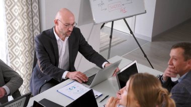 Ciddi, kendine güvenen, gözlüklü ve takım elbiseli bir adam bir masada dizüstü bilgisayar ve belgelerle oturuyor. Brifing. Bilgisayarına bakıyor ve bir şey üzerinde çalışıyor..