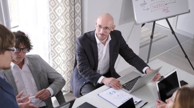 Takım elbiseli bir adam bir grup insanla aynı masada oturuyor. Elinde bir dizüstü bilgisayar tutuyor ve hoparlöre bakıyor. İş adamları önemli iş konularını tartışıyor. Sabah brifingi