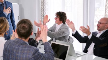 Şirketin mutlu çalışanları başarılı bir anlaşma yaptıkları için meslektaşlarını tebrik ediyor. Bir grup insan konferans salonunda ellerini çırpıyor ve birbirlerine gülümsüyor. Atmosfer olumlu ve dost canlısı.