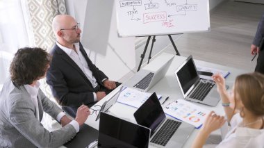 Brifingde bir iş fikri sunumu. Bir grup insan, bir beyaz tahtanın arka planında başarı grafiği ile bir masada oturuyor. Fikirleri tartışıp bir proje üzerinde çalışıyorlar..