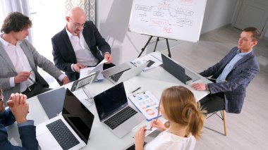 Bir grup girişimci dizüstü bilgisayar ve kağıtlarla dolu bir masada oturuyor. İş ofisinde brifing veriyorum. Uzmanlardan oluşan bir ekip iş meselelerini tartışıyor. Takım çalışması. Pazarlama, analiz ve iş stratejisi.