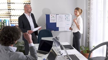 Bir adam ve bayan asistanı bir sunumla beyaz bir kurulun önünde duruyorlar. Bir kadın elinde bir işaret tutuyor ve konferans salonundaki bir grubun sorularını cevaplıyor..