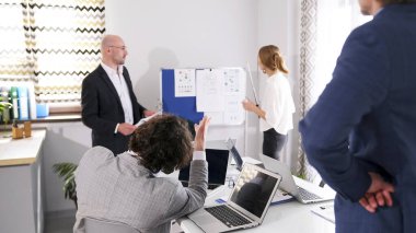 Genç bir işadamı sunum yapan bir kadın vergi danışmanına soru soruyor. Bir grup insan konferans odasında, bir adam ve asistanı beyaz bir kurulun önünde duruyor. Sahne odaklandı.