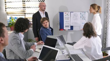 Yönetim kurulu satış müdürünün sunumunu tartışıyor. Bir grup iş adamı toplantı odasında dizüstü bilgisayarları ve kağıtlarıyla beyaz bir masada oturuyor. Sahne profesyonel ve odaklı.