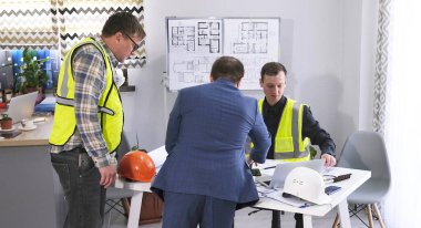 Yeşil sinyal yelekli iki inşaat işçisi ve patronları şirketin ofisinde proje planlarını tartışıyorlar. Bir güvenlik müfettişi mimarlık bürosunu kontrol ediyor. Adamlar planlara bakıyorlar.