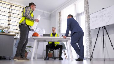 Ciddi bir müşteri bir mühendis ve inşaatçıyla bir inşaat projesinin çalışmasını tartışıyor. Mimarlık bürosundaki bir toplantıda üç adam. İkisi yeşil sinyal yelekleri giyiyor..