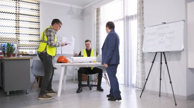 İnşaat mühendisleri çalışıyor. İki adam mimarın masasının etrafında duruyor. Mimarlık bürosunun başı çalışanlarının nasıl çalıştığını kontrol ediyor. Sahne profesyonel ve odaklı..