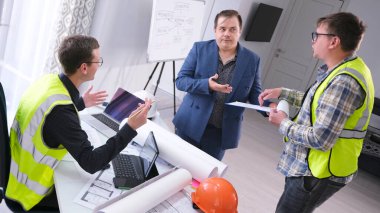 Baş mühendis ve asistanı patrondan zam istiyorlar. Üç adam mimarın ofisinde konuşuyor, ikisi yeşil sinyal yelekleri giyiyor. Sahne ciddi ve odaklanmış.