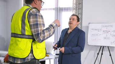 Gözlüklü bir erkek mühendis ve yeşil sinyal yeleği şirketin başkanıyla konuşuyor. İki adam proje tahminini tartışıyor. Mimarlık bürosunun ofisinde..