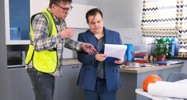 Ustabaşı inşaat projesinin detaylarını açıklıyor. İki adam mimari bir ofiste duruyor, biri yeşil bir güvenlik yeleği giyiyor. Tahmini değerlere bakıyorlar.