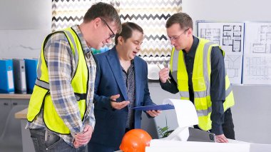 Yeşil sinyal yelekli iki işçi müşteriyle projenin çalışma aşamalarını tartışıyor. İş adamı inşaatçılara emir verir. Mimari bir nesne için tahminin onaylanması