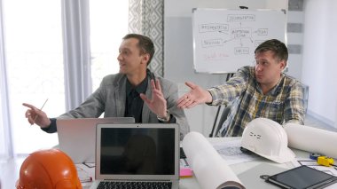 İki erkek mühendis toplantı odasında bir masada oturmuş bir inşaat projesinin detaylarını öfkeyle tartışıyorlar. Bir mimarlık şirketinde sabah planlama toplantısı..