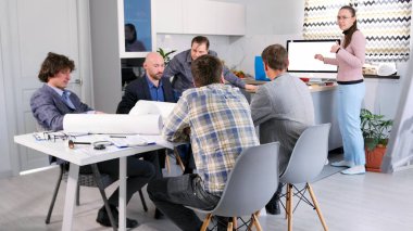 Mimarlık bürosunun yönetim kurulu yeni projenin detaylarını tartışıyor. Bir grup mühendis, inşaatçı ve mimar brifingde. Bir kadın sunum yapıyor.